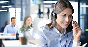 Female customer support operator with headset and smiling, with collegues at background.