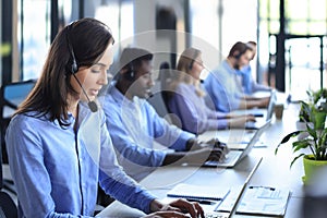 Female customer support operator with headset and smiling, with collegues at background.