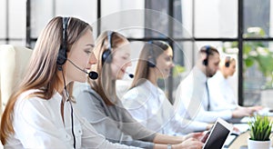 Female customer support operator with headset and smiling, with collegues at background.