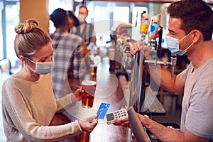 Female Customer Wearing Mask In Bar Making Contactless Payment For Drinks During Health Pandemic