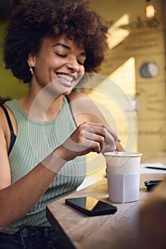 Female Customer In Window Of Cafe Stirring Hot Drink In Takeaway Cup With Wooden Stirrer