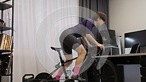 Female cyclist is pedaling out of saddle on stationary bike trainer. Indoor cycling