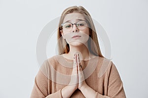 Female with dark eyes wearing eyeglasses having hopeful expression keeping her hands together asking for something