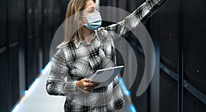 Female data center technician working inside server rack room while wearing facemask