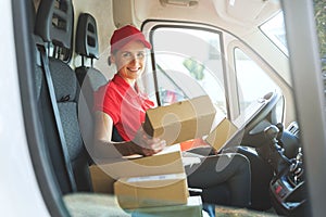 Female delivery service worker in red uniform sitting in van with boxes. smiling at camera