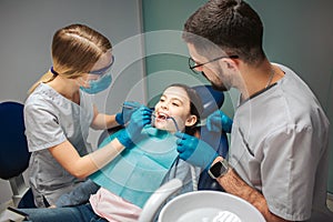 Female dentist check up girl`s teeth. Male helper stad beside. Girl sit in dental chair in room. She keep mouth opened.