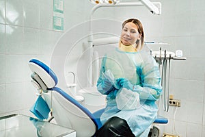 A female dentist with a medical mask and rubber gloves in her hands sits in her office