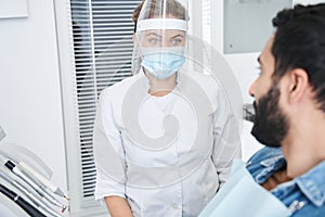 Female dentist in protective mask talking with her male patient