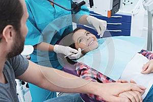 A female dentist is treating a girl. She is sitting on the dental chair. She is afraid and her father holds her hand.