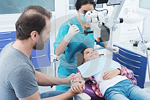 A female dentist is treating a girl. She is sitting on the dental chair. She is afraid and her father holds her hand.