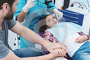 A female dentist is treating a girl. She is sitting on the dental chair. She is afraid and her father holds her hand.