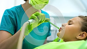 Female dentist using saliva ejector, teenage girl sitting in chair frowning