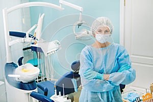 A female dentist wearing a medical mask and rubber gloves poses for the camera and folds her arms in her office