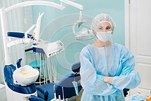 A female dentist wearing a medical mask and rubber gloves poses for the camera and folds her arms in her office