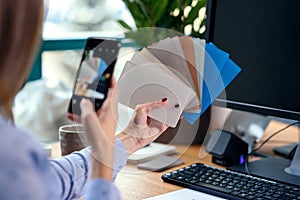 Female designer in office working with colour samples. Woman at workplace makes a photo of color palette on smartphone.