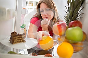 Female on diet in dilemma whether to eat chocolate cake or orange from fridge