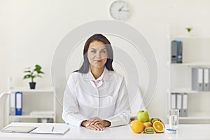 Female dietician sitting at office desk with fruit and measuring tape looking at camera