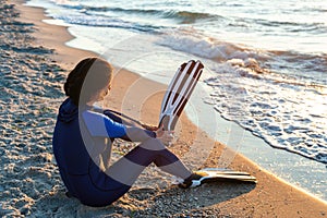 Female diver in blue wetsuit puts on flippers sitting on the beach