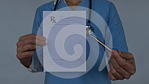 A female doctor in a blue uniform is holding a blank prescription pad and pen. She is looking at the camera with a