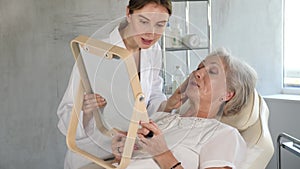 Female doctor cosmetologist stand with woman looking mirror behind in the doctors office.