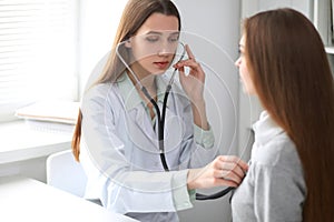 Female doctor examining her patient with stethoscope while sitting at the table near the window in hospital. Physician