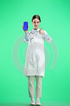 A female doctor, full-length, on a green background, shows a phone