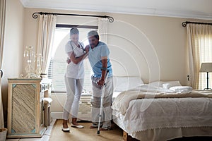 Female doctor helping senior man to walk with crutches in bedroom