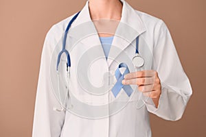 Female doctor holding blue ribbon on color background, closeup. Prostate cancer awareness concept