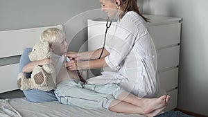 A female doctor is listening with a stethoscope to a cute blond boy at his home