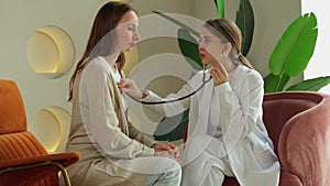 Female doctor listens to the lungs of a young woman sitting on the couch with a stethoscope. A cardiologist checks the