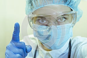 A female doctor in a medical mask, glasses, and gloves sternly hold up her index finger, a sign of advice to be careful.