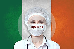 Female doctor in medical mask with the inscription COVID-19 on a blurred background of the flag of  Ireland. Pandemic virus COVID-