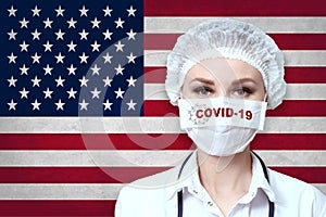 Female doctor in medical mask with the inscription COVID-19 on a blurred background of the flag of USA flag. Pandemic virus COVID-
