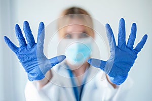 Female doctor with a medical mask showing hands to camera while wearing a blue nitrile gloves over white background