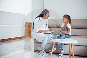 Female doctor with notepad sits in the clinic with little girl
