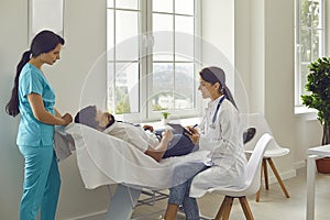 Female doctor and a nurse examine a patient lying on a couch in a hospital room.