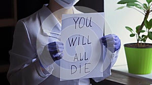 Female doctor in protective mask and suit slowly showing sheet of paper with text: you will all die. Virologist lift up