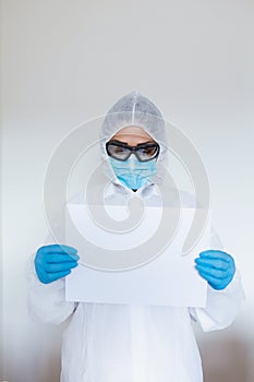 Female doctor with protective workwear holding empty cardboard while standing at clinic and looking at camera. Copy