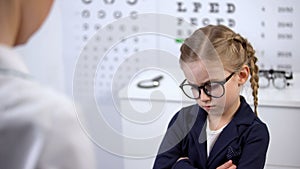 Female doctor putting glasses on disgruntled girl, child feels insecure, upset