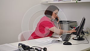 A female doctor in a red medical suit looks through documents lying on the table. A woman picks up the phone, dials a