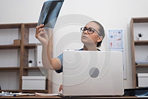 female doctor scrutinizes patient X-ray scan, concurrently checking test results on her laptop