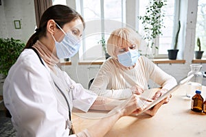 Female doctor and senior woman, patient wearing face mask, having appointment in office. Checkup and treatment after