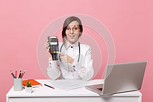 Female doctor sit at desk work on computer with medical document credit card in hospital isolated on pastel pink wall