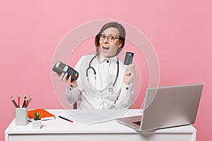 Female doctor sit at desk work on computer with medical document credit card in hospital isolated on pastel pink wall