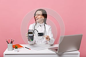 Female doctor sit at desk work on computer with medical document credit card in hospital isolated on pastel pink wall