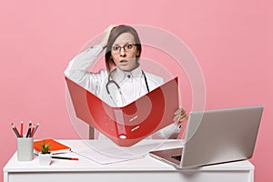Female doctor sit at desk work on computer with medical document hold folder in hospital isolated on pastel pink wall