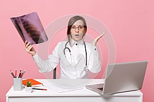 Female doctor sit at desk work on computer with medical document hold X-ray in hospital isolated on pastel pink wall