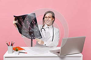 Female doctor sit at desk work on computer with medical document hold x-ray in hospital isolated on pastel pink wall