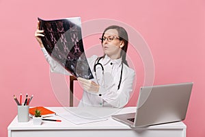 Female doctor sit at desk work on computer with medical document hold x-ray in hospital isolated on pastel pink wall