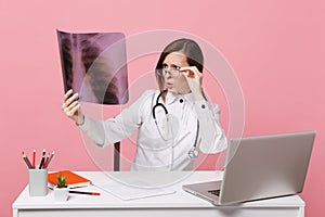 Female doctor sit at desk work on computer with medical document hold X-ray in hospital isolated on pastel pink wall